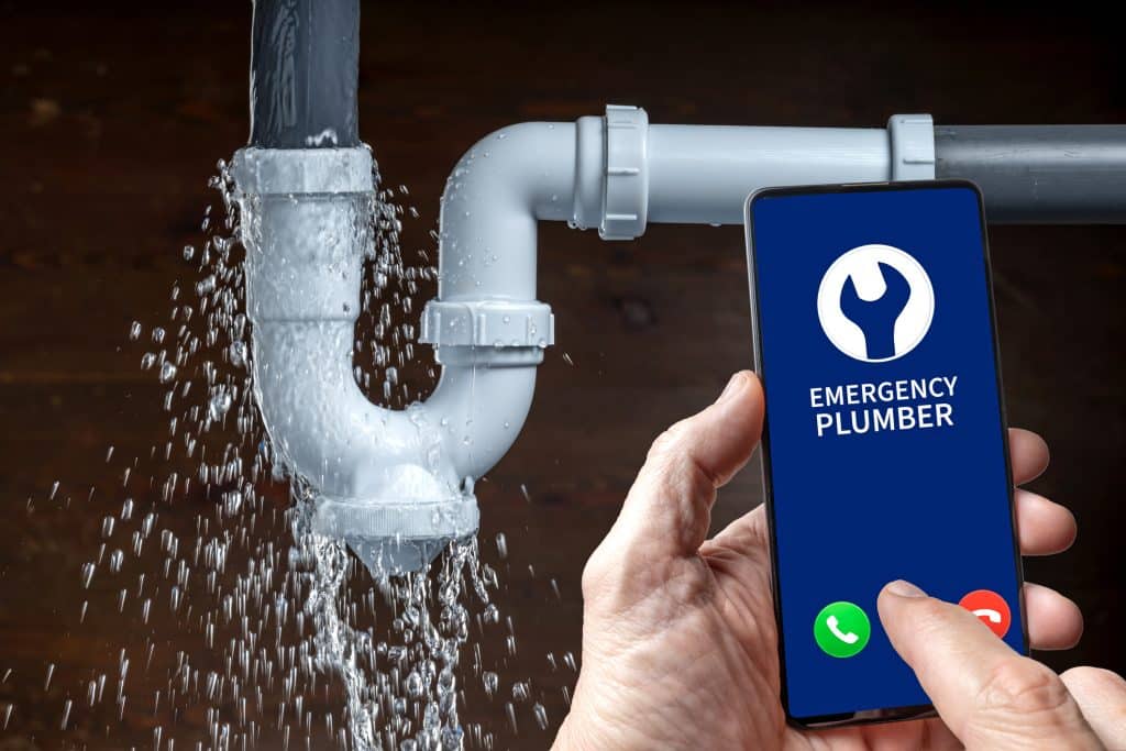 Hands hold a mobile phone while dialling a plumber with a leaky pipe in the background.
